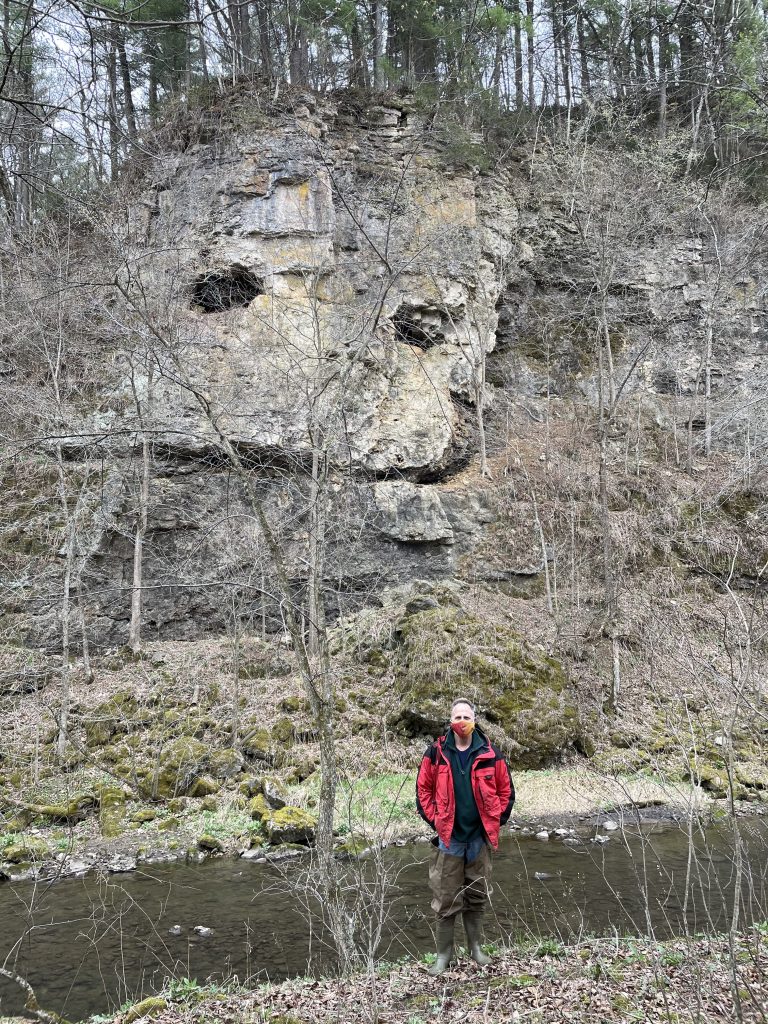 Tony Runkel standing next to an outcrop of Oneota dolomite in the Whitewater Wildlife Management Area in southeastern Minnesota. Tony is wearing a red coat, red mask, and waders.